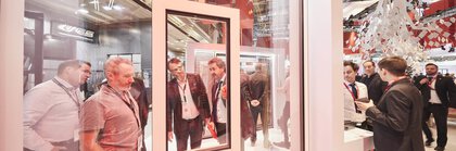 Several men stand behind a glass wall with a recessed glass window at a stand at the BAU trade fair and look at the construction.
