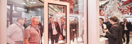 Mehrere Männer stehen hinter einer gläsernen Wand mit einem eingelassenen gläsernen Fenster an einem Stand auf der BAU-Messe und betrachten die Konstruktion.