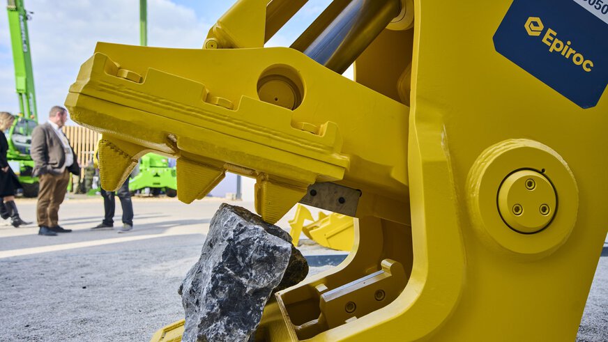 Yellow Epiroc grapple tool holding a rock; people and green construction machines visible in the background at a trade show.