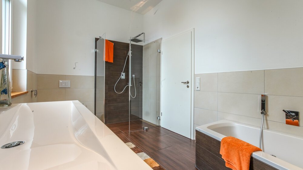 Modernes Badezimmer mit Badewanne, Glasdusche und orangefarbenen Handtüchern auf Ständern. Helle Beleuchtung und neutrales Farbschema.