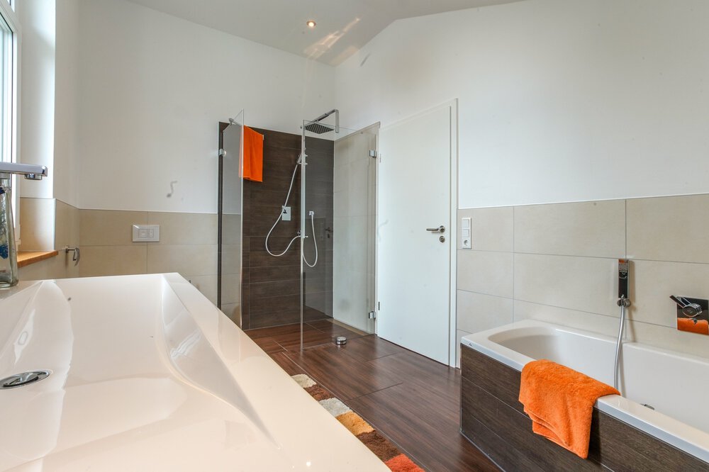 Modernes Badezimmer mit Badewanne, Glasdusche und orangefarbenen Handtüchern auf Ständern. Helle Beleuchtung und neutrales Farbschema.