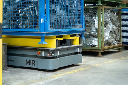 Autonomous robot transporting a heavy metal cage full of parts in a warehouse.