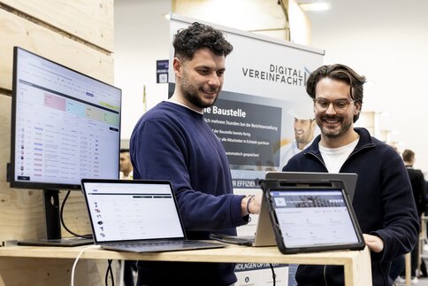 Zwei Männer lächeln und arbeiten an Laptops an einem technischen Stand mit Bildschirmen, auf denen Software angezeigt wird.