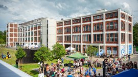 Eine Menschenmenge versammelt sich bei einer Freiluftveranstaltung in der Nähe eines großen, modernen Gebäudes aus Ziegeln und weißer Farbe unter einem bewölkten Himmel.