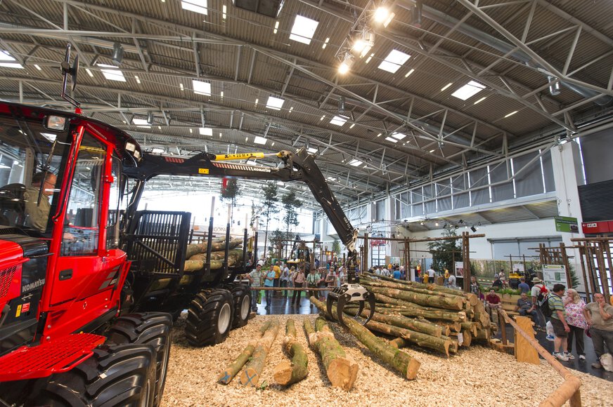 Exhibition hall at INTERFORST: A red forestry machine with a black grapple lifts a tree trunk in front of an audience, other trunks on a pile.