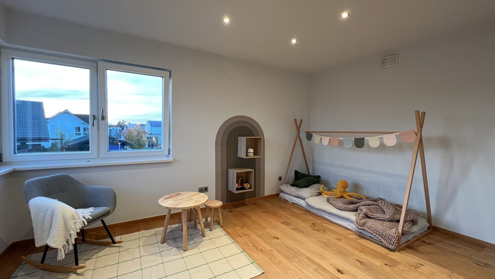 Minimalistisches Kinderzimmer mit einem Bodenbett, Spielzeug, einem kleinen Tisch, einem Schaukelstuhl und einem großen Fenster.