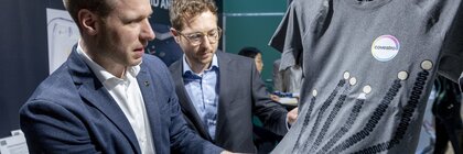 Two men in suits are looking at a T-shirt with printed electronics and Covestro logo at a trade show.