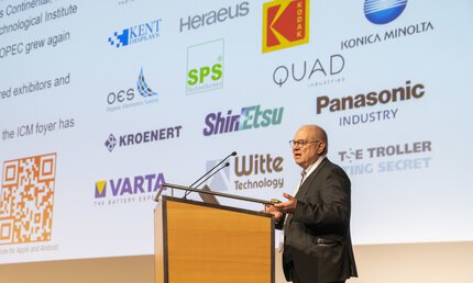 A speaker on a podium presents in front of a screen on which various company logos are displayed.