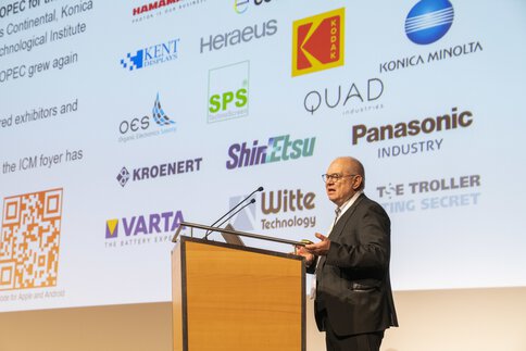 A speaker on a podium presents in front of a screen on which various company logos are displayed.