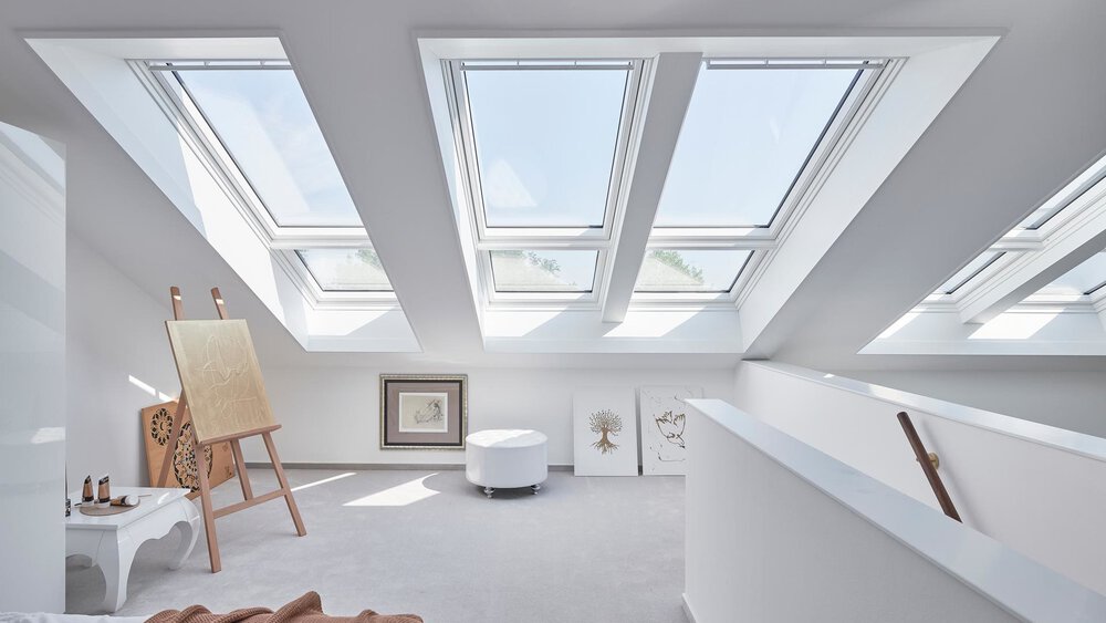 Helles, modernes Dachgeschoss mit großen Oberlichtern, weißen Wänden, Holzmöbeln und einem kleinen Bücherregal.