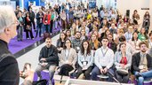 View from the stage into the audience, which is concentrating on a presentation. Predominantly young people sit and stand in an exhibition hall, with exhibition stands visible in the background.