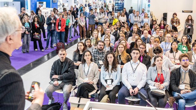 View from the stage into the audience, which is concentrating on a presentation. Predominantly young people sit and stand in an exhibition hall, with exhibition stands visible in the background.