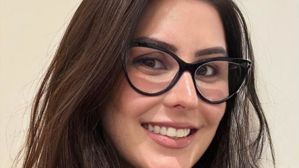 A smiling woman with striking black glasses and long dark hair is wearing a black top and a necklace.