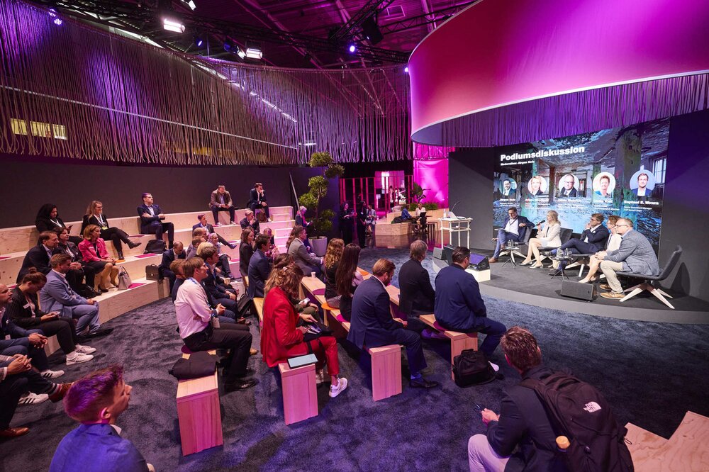 Five people on stage at a panel discussion in front of a seated audience in a purple-lit carpeted area.
