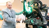 A man in a grey suit operates a large robotic device with green lights at the trade fair.