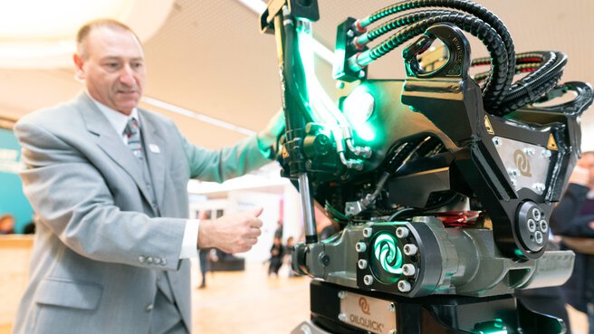A man in a grey suit operates a large robotic device with green lights at the trade fair.