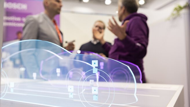 Three people discussing at a trade fair stand. Three illuminated silhouettes of cars cut out of Plexiglas are displayed in the foreground.