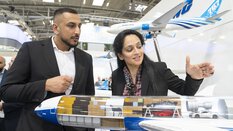Ein Mann und eine Frau in Businesskleidung betrachten das Innenleben eines Flugzeugmodells mit durchsichtigem Frachtraum an einem Messestand.