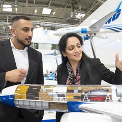 Ein Mann und eine Frau in Businesskleidung betrachten das Innenleben eines Flugzeugmodells mit durchsichtigem Frachtraum an einem Messestand.