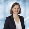 Lächelnde Frau mit glatten braunen Haaren in einem dunklen Blazer und einer weißen Bluse.