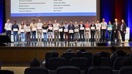 A group of people on stage hold certificates in their hands. A list of names can be seen on the screen behind them.