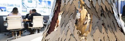 Nahaufnahme einer baumähnlichen Skulptur in einem Büro. Im Hintergrund sitzen Menschen an einem Tisch.