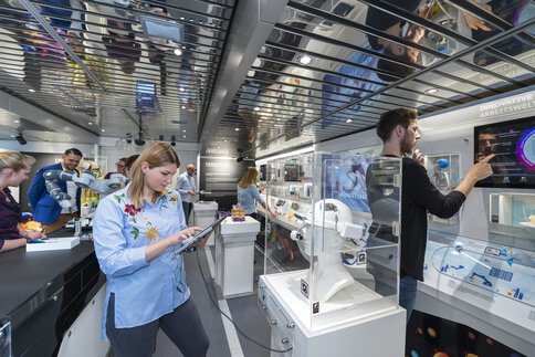 Mehrere Besucher stehen in einem niedrigen Ausstellungsraum mit spiegelnder Decke und interagieren mit Displays und technischen Exponaten.