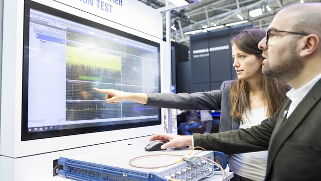 Zwei Personen stehen an einem Stand auf der productronica und blicken auf einen großen Monitor mit Diagrammen. Die Frau deutet auf etwas, der Mann greift zur Maus.