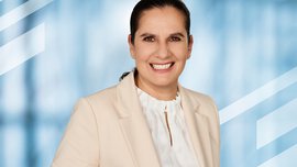 Portrait of Susanne Grödl, Exhibition Director of analytica Munich, smiling in front of a light blue background, wearing a cream blazer and a white blouse.