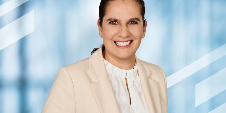 Portrait of Susanne Grödl, Exhibition Director of analytica Munich, smiling in front of a light blue background, wearing a cream blazer and a white blouse.