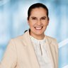 Portrait of Susanne Grödl, Exhibition Director of analytica Munich, smiling in front of a light blue background, wearing a cream blazer and a white blouse.