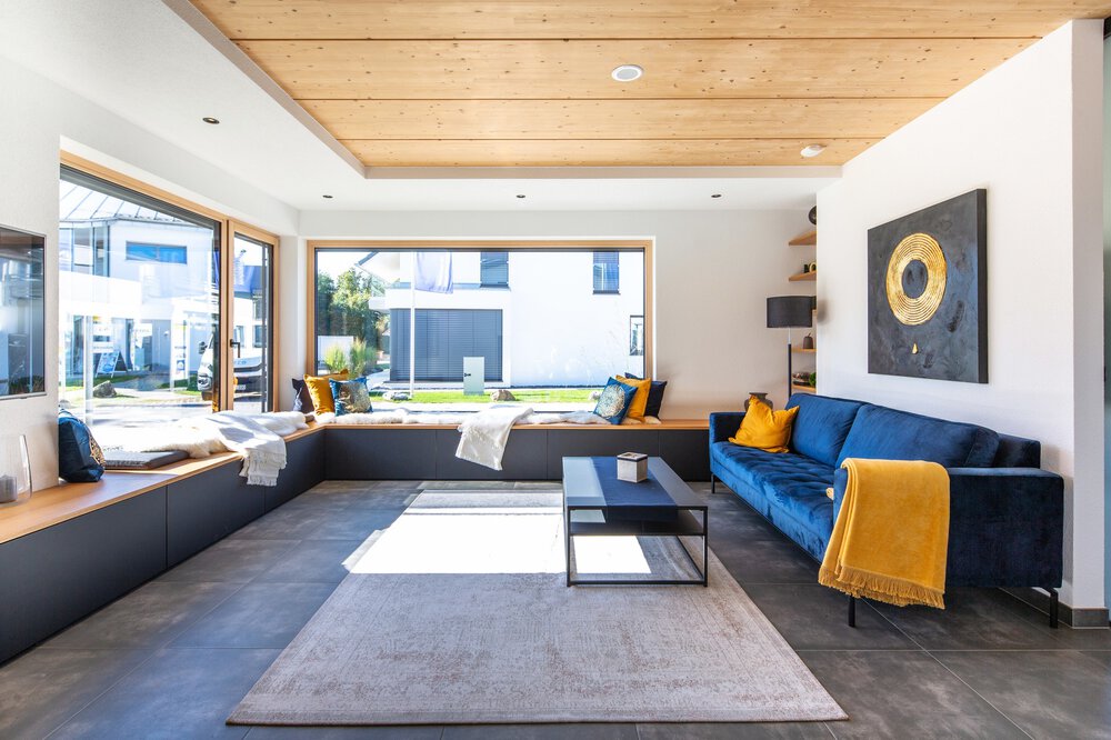 Modernes Wohnzimmer mit blauem Sofa, gelben Kissen, großen Fenstern und einem schwarzen Couchtisch auf einem hellen Teppich.