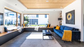 Modernes Wohnzimmer mit blauem Sofa, gelben Kissen, großen Fenstern und einem schwarzen Couchtisch auf einem hellen Teppich.