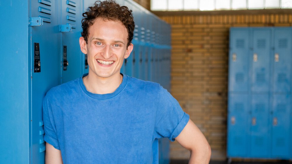 Ein lächelnder Mann mit kurzen gelockten Haaren und in einem blauen T-Shirt lehnt in einer Umkleidekabine an einer Reihe von blauen Spinden.