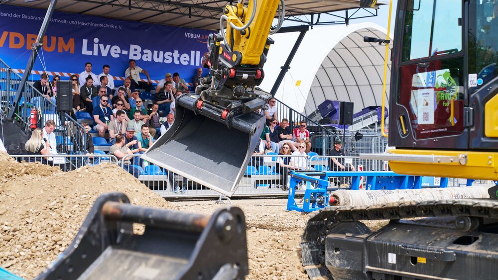 Live demonstration at the VDBUM live construction site: A yellow excavator moving earth in front of a spectator grandstand in the outdoor area.