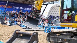 Live demonstration at the VDBUM live construction site: A yellow excavator moving earth in front of a spectator grandstand in the outdoor area.