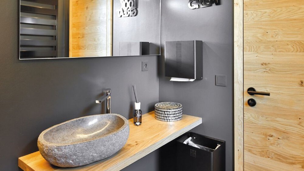 Modernes Badezimmer mit Steinwaschbecken auf einer Holzplatte vor einem großem Spiegel und grauen Wänden.