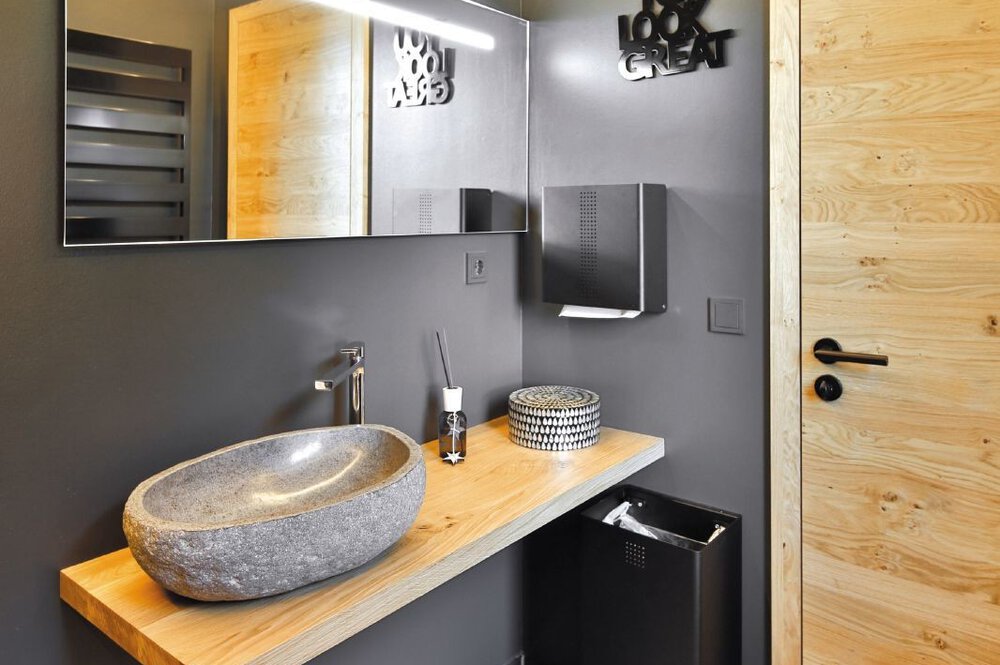 Modernes Badezimmer mit Steinwaschbecken auf einer Holzplatte vor einem großem Spiegel und grauen Wänden.