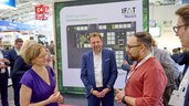 Three people in conversation: woman in purple dress, man in blue suit, and bearded man in red shirt standing in front of a layout map of the Start-up Area at IFAT Munich.