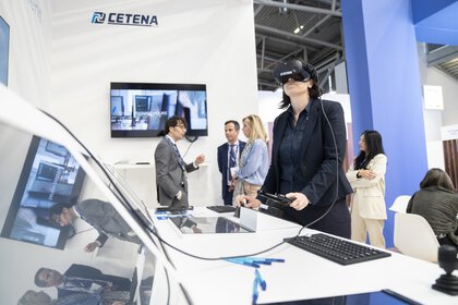 Eine Frau, die ein VR-Headset trägt und zwei Bedienelemente an einem Stand der transport logistic testet, während sich hinter ihr eine Gruppe von Leuten unterhält.