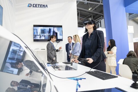 Eine Frau, die ein VR-Headset trägt und zwei Bedienelemente an einem Stand der transport logistic testet, während sich hinter ihr eine Gruppe von Leuten unterhält.