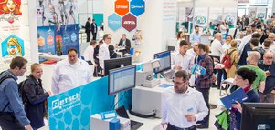 Messestand mit Personen an Computern und im Gespräch, im Hintergrund Banner mit Logos.