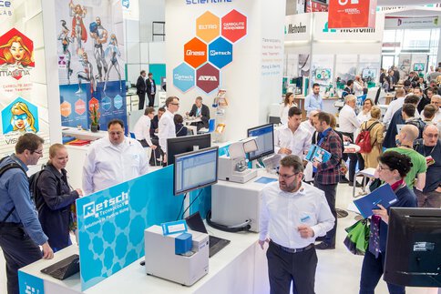 Messestand mit Personen an Computern und im Gespräch, im Hintergrund Banner mit Logos.