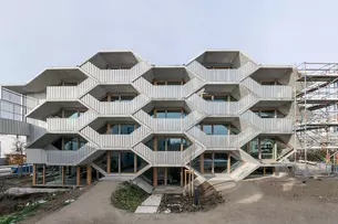 Geometrische Gebäudefassade mit sechseckigen Fenstern im Bau, umgeben von einem Gerüst und einem Feldweg.