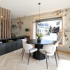 Modernes Esszimmer mit rundem schwarzen Tisch, vier weißen Stühlen, geblümter Tapete und großem Fenster.