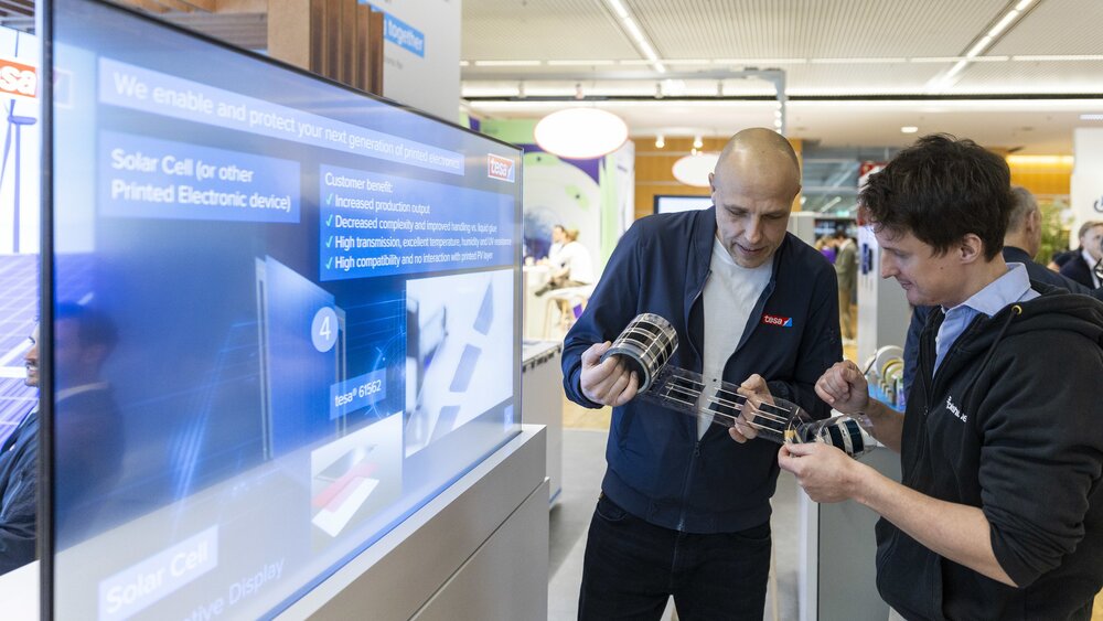 Zwei Männer stehen vor einer illuminierten blauen Informationstafel an einem Stand auf der LOPEC und entrollen eine Folie mit flexiblen Leiterplatinen.