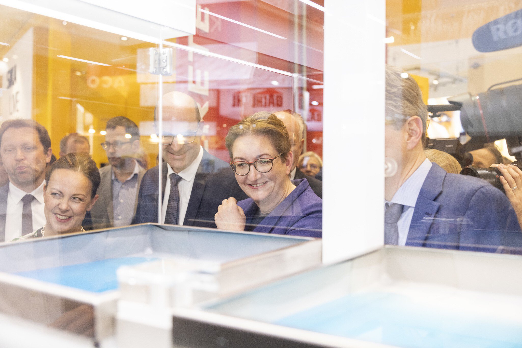 A group of smiling people in business attire look into a display case at a BAU trade fair stand, while members of the press capture the action.
