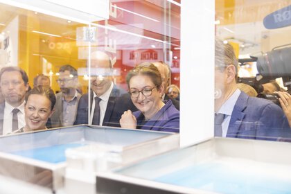 Eine Gruppe von lächelnden Menschen in Businesskleidung blickt in eine Vitrine auf der BAU-Messe, während Presseleute das Geschehen festhalten.