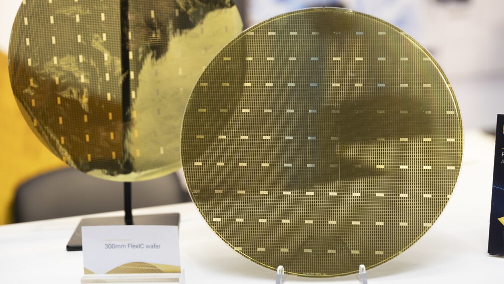 Nahaufnahmen zweier goldener Leiterplatinen in runder Form. Davor steht ein Schild mit der Bezeichnung „300mm FlexIC wafer“.