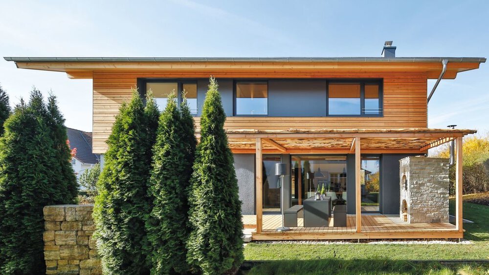 Modernes zweistöckiges Haus mit Holzverkleidung, großen Fenstern und einer Veranda, umgeben von grünen Bäumen und einer Steinmauer.